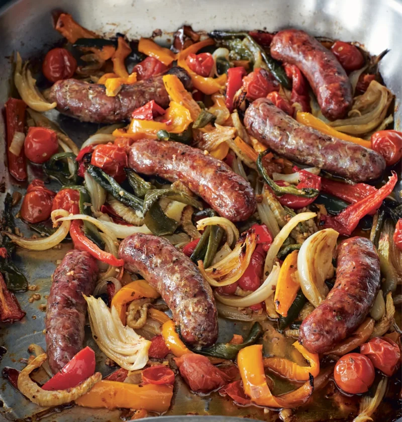 Ina Garten roasted sausages, peppers & onions in a metal roasting pan with red and orange peppers, onions, fennel and blistered cherry tomatoes