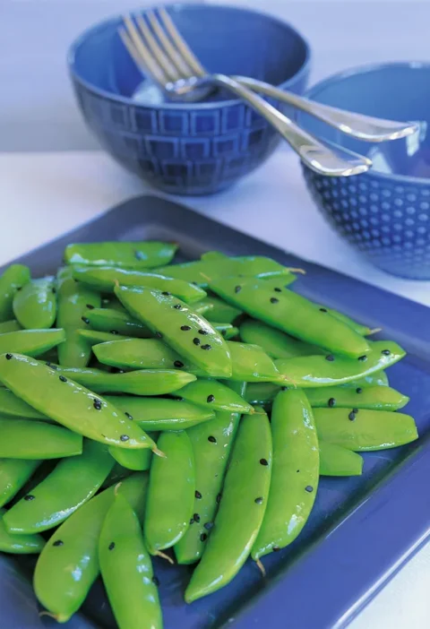 Ina Garten sugar snap peas with sesame piled on a navy blue platter, glossy green pods scattered with black sesame seeds