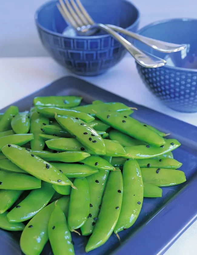 Ina Garten sugar snap peas with sesame piled on a navy blue platter, glossy green pods scattered with black sesame seeds