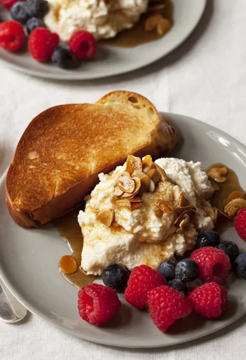 A plate of creamy breakfast ricotta topped with butter-toasted almonds, maple syrup, and fresh mixed berries served with brioche