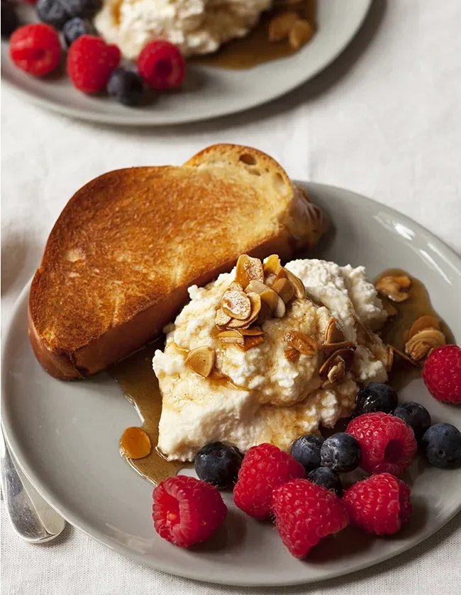 A plate of creamy breakfast ricotta topped with butter-toasted almonds, maple syrup, and fresh mixed berries served with brioche