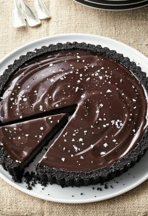 A dark chocolate tart with glossy ganache topping and flaked sea salt on a serving plate, an Ina Garten dark chocolate tart recipe