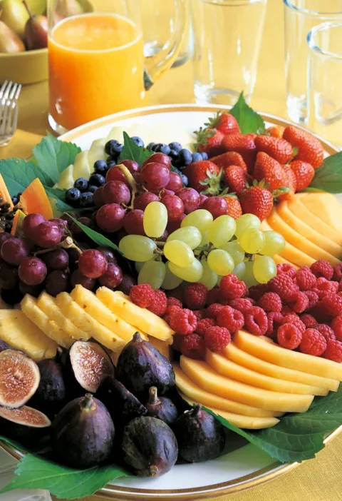 A colorful fresh fruit platter with sliced melon, golden pineapple, grapes, figs, berries, and papaya arranged on a serving platter