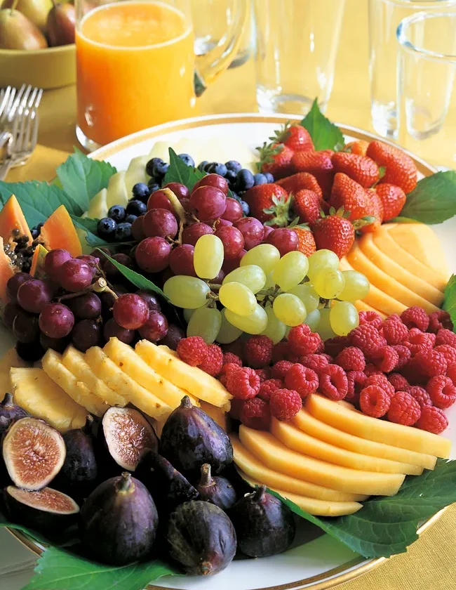 A colorful fresh fruit platter with sliced melon, golden pineapple, grapes, figs, berries, and papaya arranged on a serving platter