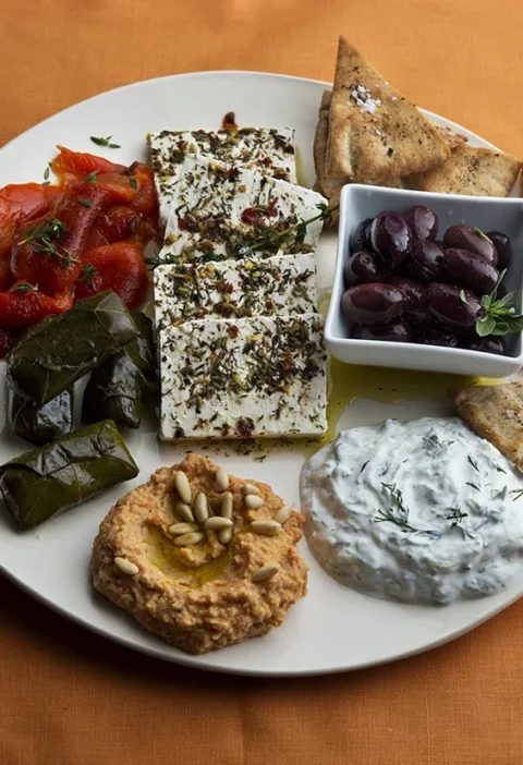 Greek mezze platter with thyme roasted red peppers, hummus, herbed feta, tzatziki, olives, grape leaves, and pita triangles arranged on a serving board