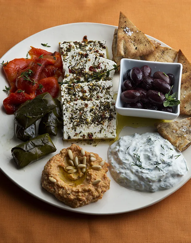Greek mezze platter with thyme roasted red peppers, hummus, herbed feta, tzatziki, olives, grape leaves, and pita triangles arranged on a serving board