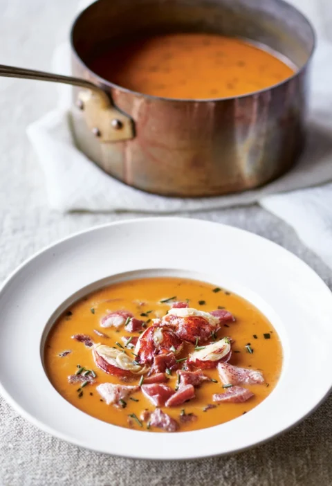 Ina Garten Maine lobster stew in a white bowl with chunks of red lobster in golden saffron cream broth topped with chives and a copper saucepan behind