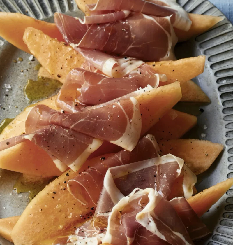 A plate of ripe cantaloupe slices with Italian prosciutto di Parma rippled on top, served with lemon wedges for Ina Garten melon and prosciutto recipe