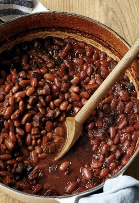 A Dutch oven filled with rich, mahogany-colored molasses baked beans with chunks of pork belly and bacon