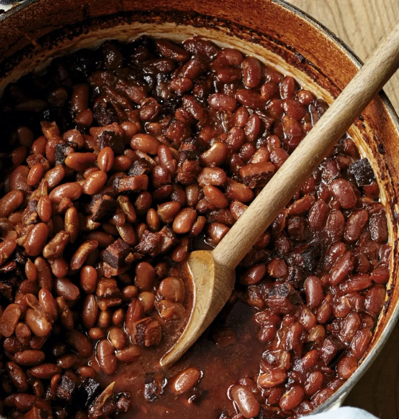 A Dutch oven filled with rich, mahogany-colored molasses baked beans with chunks of pork belly and bacon
