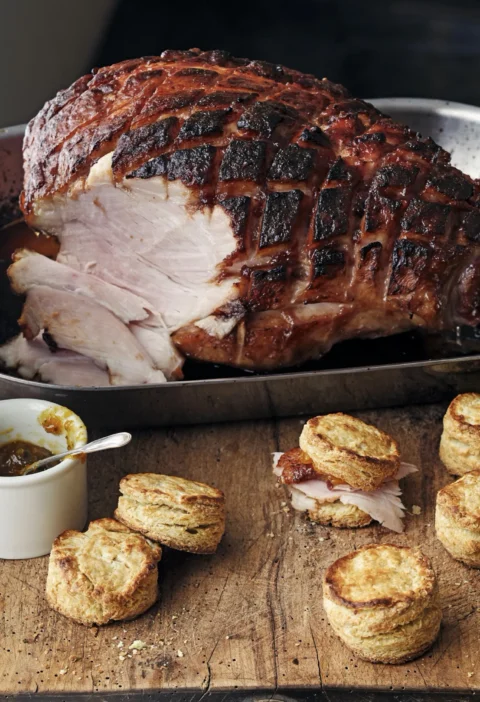 A golden-brown bone-in smoked ham coated in a sticky orange marmalade glaze, scored in a diamond pattern, in a roasting pan
