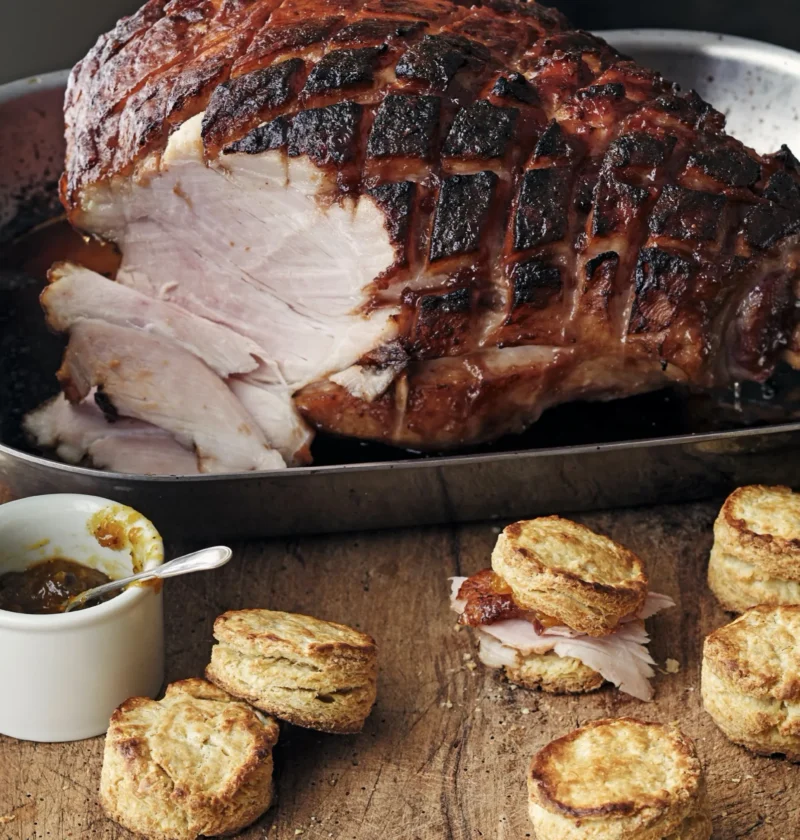 A golden-brown bone-in smoked ham coated in a sticky orange marmalade glaze, scored in a diamond pattern, in a roasting pan