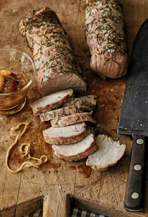 Sliced rosemary roasted pork tenderloins on a cutting board with fresh rosemary sprigs and mango chutney, an ina garten rosemary roasted pork tenderloins recipe