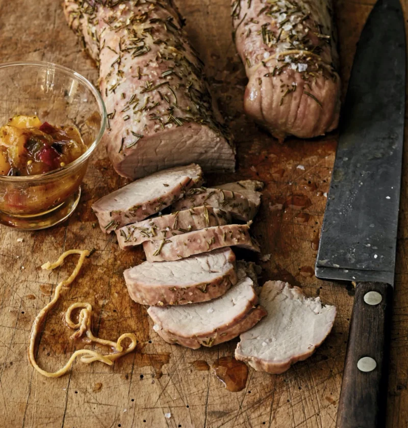 Sliced rosemary roasted pork tenderloins on a cutting board with fresh rosemary sprigs and mango chutney, an ina garten rosemary roasted pork tenderloins recipe