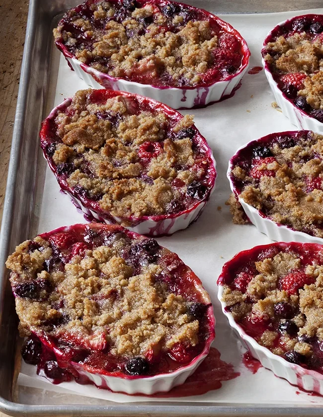 Individual tri-berry crumbles in white ramekins with bubbly blueberry, raspberry, and strawberry filling topped with golden oat crumble, an ina garten tri-berry crumbles recipe
