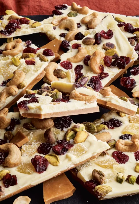 Pieces of white chocolate toffee topped with cashews, pistachios, crystallized ginger, and dried cranberries on a serving board, Ina Garten white chocolate toffee recipe