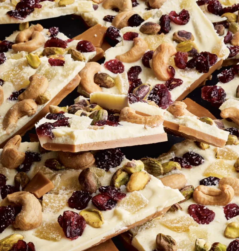 Pieces of white chocolate toffee topped with cashews, pistachios, crystallized ginger, and dried cranberries on a serving board, Ina Garten white chocolate toffee recipe
