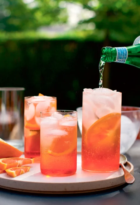 Ina Garten Aperol spritzer in tall glasses with ice and orange slices while sparkling water is poured in from a green bottle