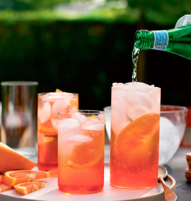 Ina Garten Aperol spritzer in tall glasses with ice and orange slices while sparkling water is poured in from a green bottle