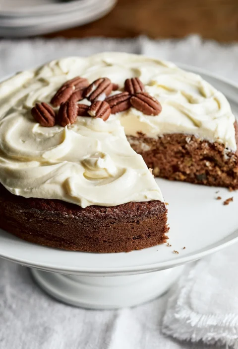 Ina Garten applesauce cake with bourbon raisins topped with swirled cream cheese frosting and pecan halves on a white cake stand, with a slice removed