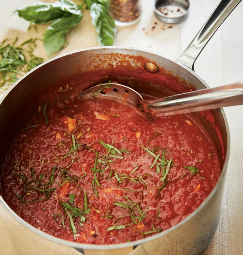 Ina Garten arrabbiata sauce simmering in a stainless steel pot with fresh basil and red pepper flakes beside it
