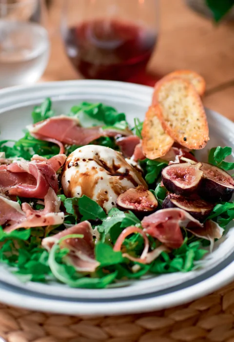 Ina Garten arugula with prosciutto and burrata topped with halved fresh figs and served with garlic toasts on a white plate