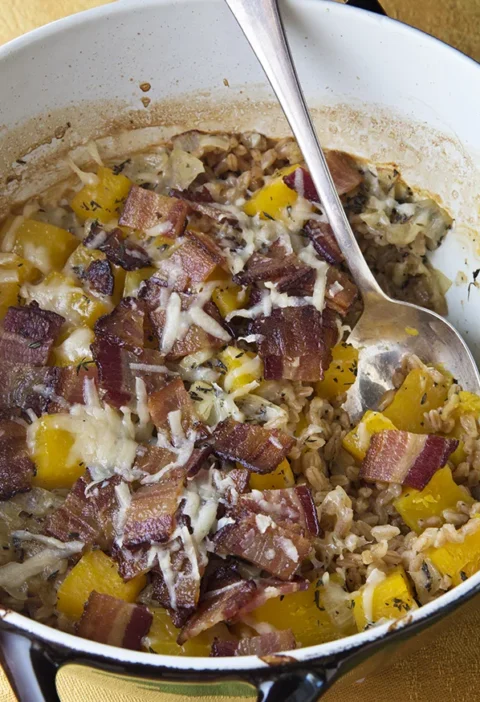 Ina Garten baked farro and butternut squash with bacon and melted Parmesan in a Dutch oven with a serving spoon