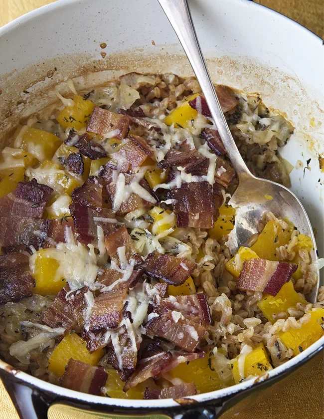 Ina Garten baked farro and butternut squash with bacon and melted Parmesan in a Dutch oven with a serving spoon