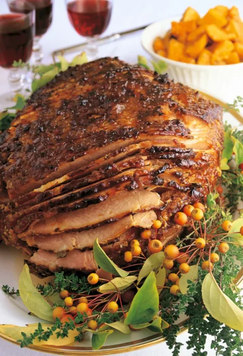 Ina Garten baked Virginia ham, spiral-cut and glazed dark brown on a platter with herbs, crab apples and a bowl of roasted squash