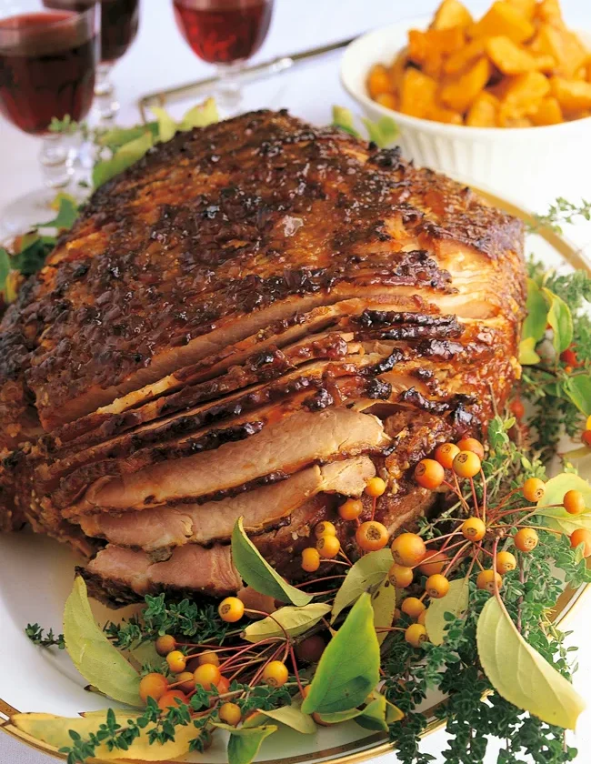 Ina Garten baked Virginia ham, spiral-cut and glazed dark brown on a platter with herbs, crab apples and a bowl of roasted squash