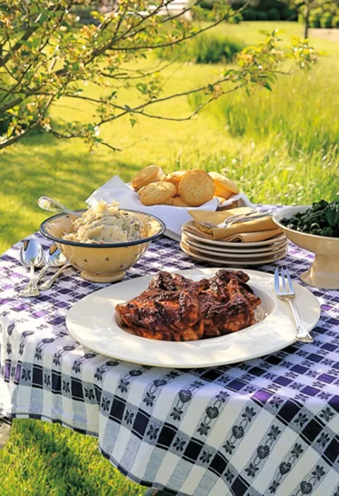 Ina Garten barbecued chicken on a white platter at an outdoor table with mashed potatoes, biscuits and greens