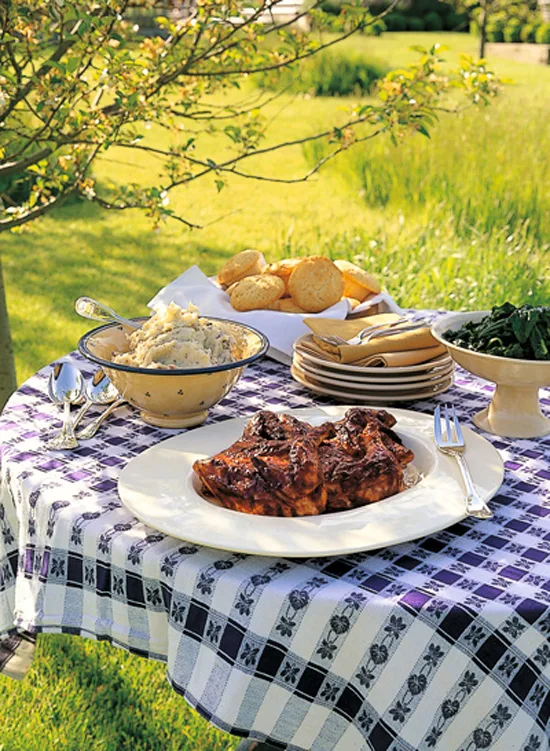 Ina Garten barbecued chicken on a white platter at an outdoor table with mashed potatoes, biscuits and greens