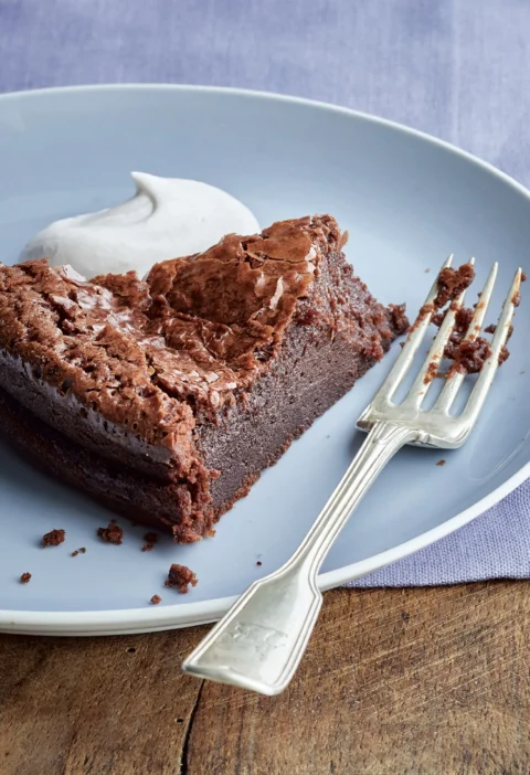 Slice of Ina Garten bittersweet chocolate cake with a crackly top and a dollop of whipped cream on a blue plate with a silver fork