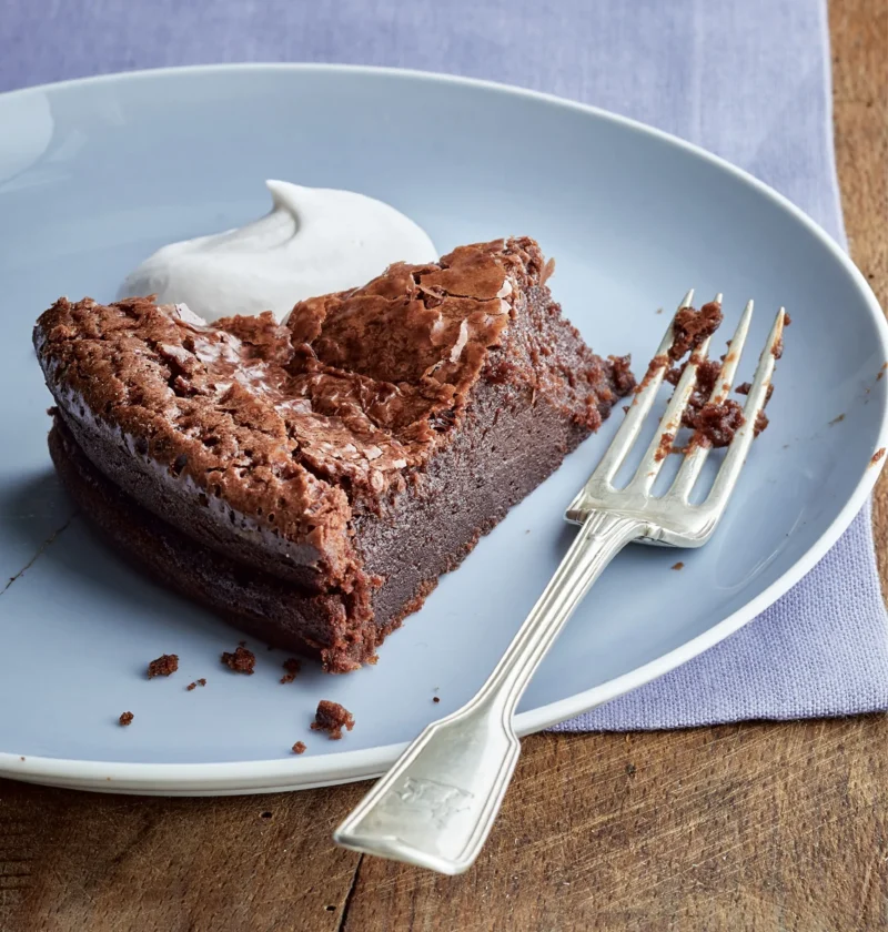 Slice of Ina Garten bittersweet chocolate cake with a crackly top and a dollop of whipped cream on a blue plate with a silver fork