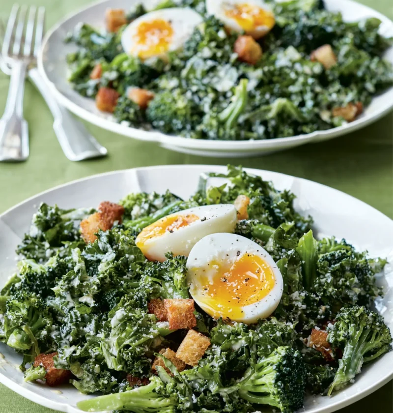 Ina Garten broccoli & kale salad with halved soft-boiled eggs, croutons and Caesar dressing on two white plates