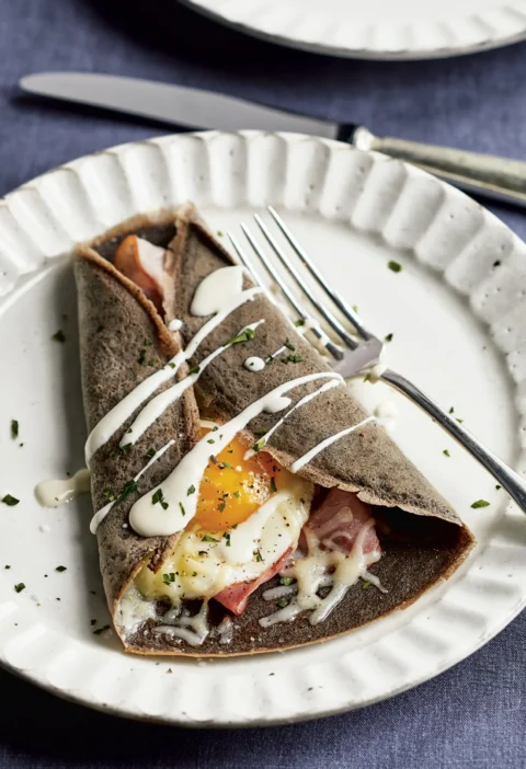Ina Garten buckwheat crêpes complète folded around ham, melted Gruyère and a fried egg with crème fraîche drizzle on a white plate