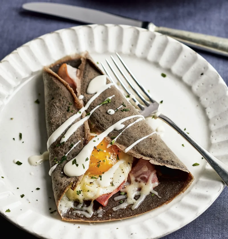 Ina Garten buckwheat crêpes complète folded around ham, melted Gruyère and a fried egg with crème fraîche drizzle on a white plate