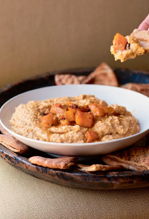 Ina Garten butternut squash hummus in a white bowl topped with roasted squash cubes, with toasted pita wedges around it