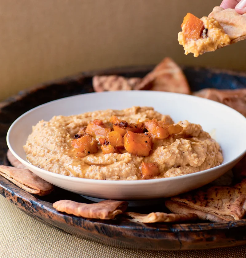 Ina Garten butternut squash hummus in a white bowl topped with roasted squash cubes, with toasted pita wedges around it