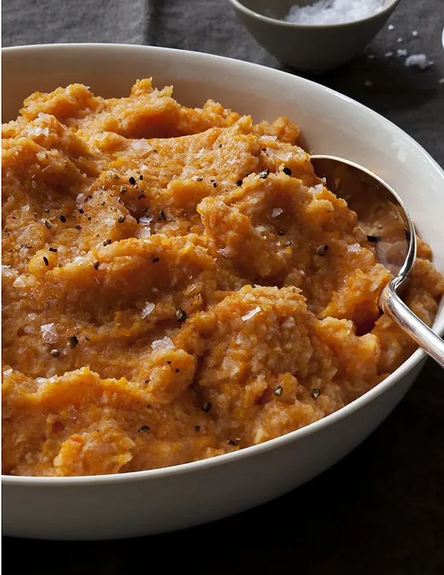 Ina Garten carrot and cauliflower purée in a white bowl with a serving spoon, topped with flaky salt and black pepper
