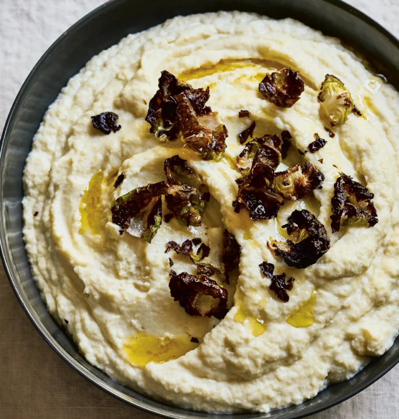 Ina Garten celery root & cauliflower purée in a dark bowl topped with crispy roasted Brussels sprout leaves and a drizzle of olive oil