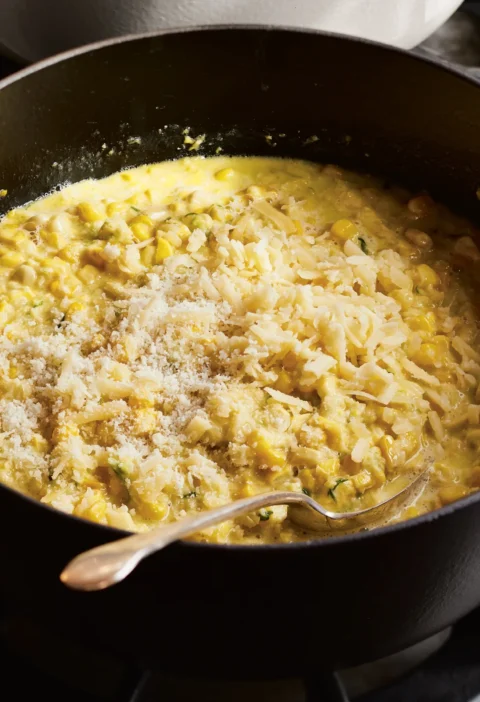 Ina Garten cheddar & scallion creamed corn in a black pot topped with grated Cheddar and Parmesan with a spoon resting in it