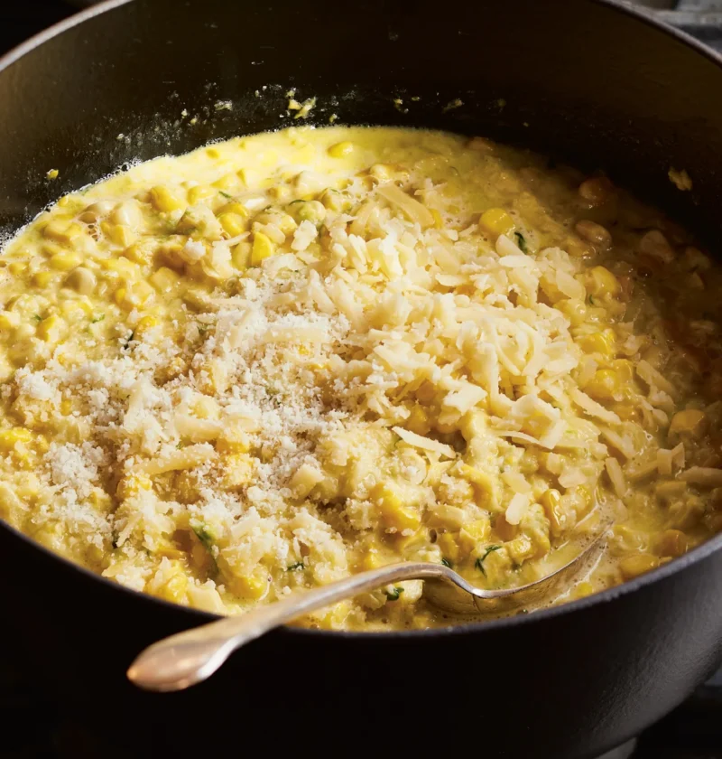 Ina Garten cheddar & scallion creamed corn in a black pot topped with grated Cheddar and Parmesan with a spoon resting in it