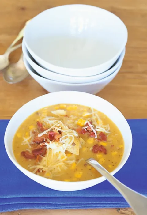 Ina Garten cheddar corn chowder in a white bowl topped with shredded cheese and crisp bacon on a blue napkin