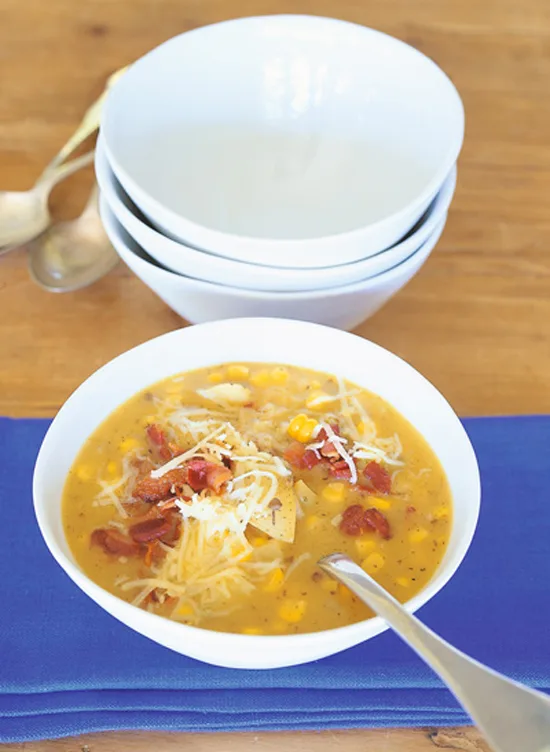 Ina Garten cheddar corn chowder in a white bowl topped with shredded cheese and crisp bacon on a blue napkin