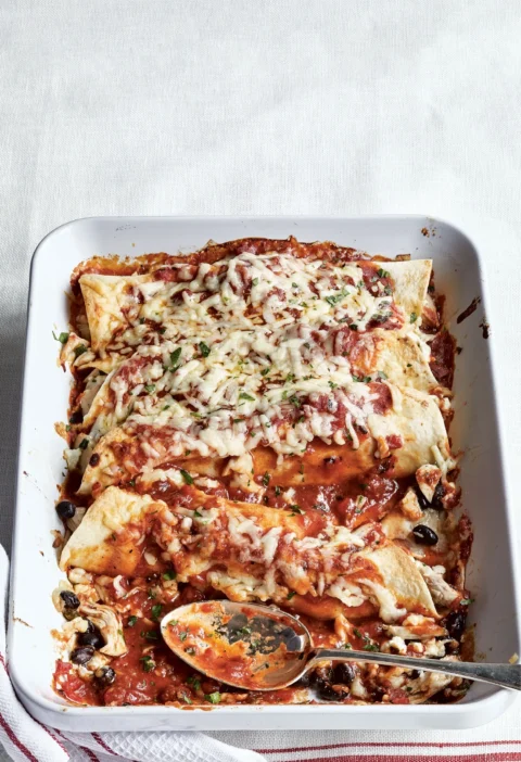 Ina Garten cheesy chicken enchiladas in a white baking dish topped with melted cheddar, red sauce and parsley with a serving spoon