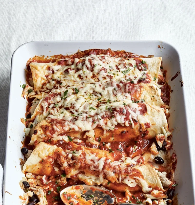Ina Garten cheesy chicken enchiladas in a white baking dish topped with melted cheddar, red sauce and parsley with a serving spoon