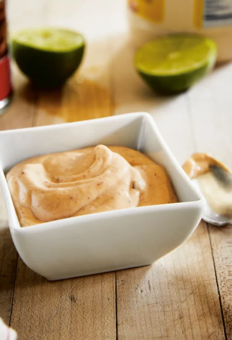 Ina Garten chipotle mayonnaise swirled in a white square bowl next to lime halves and a spoon on a wooden board