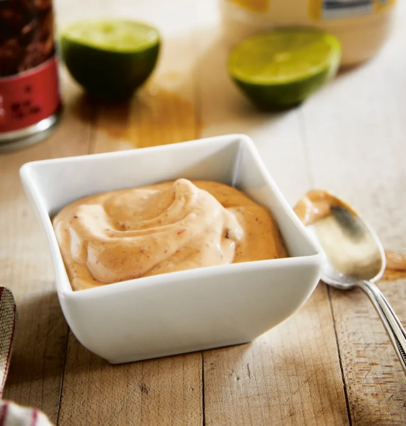 Ina Garten chipotle mayonnaise swirled in a white square bowl next to lime halves and a spoon on a wooden board