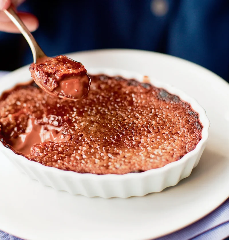 Ina Garten chocolate crème brûlée in a white fluted ramekin with a caramelized sugar top and a spoon lifting out a bite of creamy chocolate custard